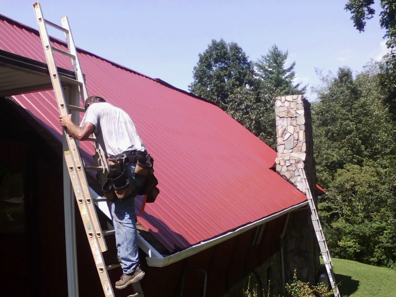 Roofer on a ladder beside a red metal roof and stone chimney — Soffit & Fascia Repair by Roofing Brothers in Rothsay